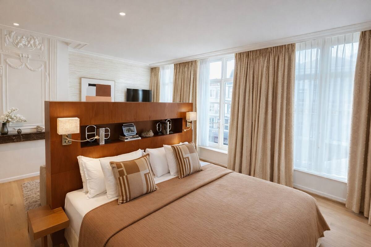 A hotel room with a large bed, a wooden headboard, a flat-screen TV, a bedside table, and a window with curtains.
