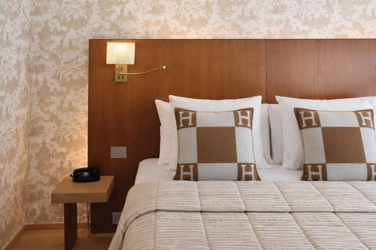 A hotel room with a wooden headboard, a bedside lamp, a telephone, and a bed with pillows featuring a checkered pattern.