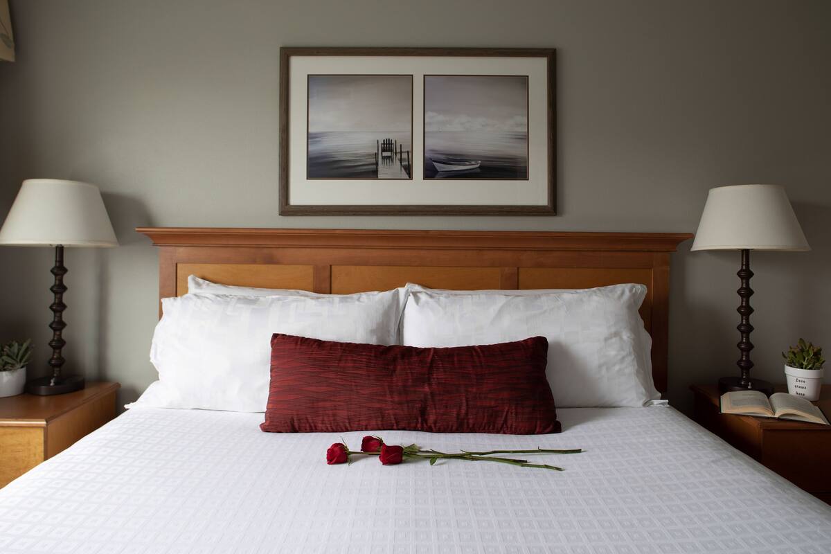 A neatly made bed with white and red pillows, a framed picture above, and a rose on the bed.