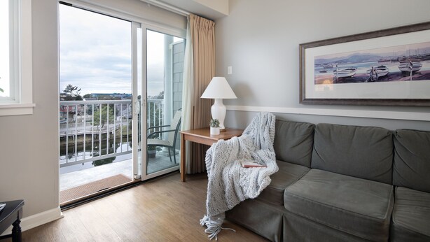 A modern living room with a grey sofa, a small table with a lamp, a framed picture on the wall, and a sliding glass door leading to a balcony with a view of buildings and water.