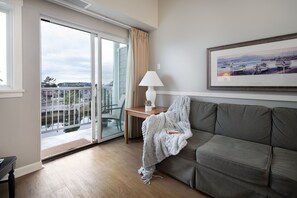 Una sala de estar moderna con un sofá gris, una mesita con una lámpara, un cuadro enmarcado en la pared y un ventanal corredero que da a un balcón con vistas a edificios y al agua.