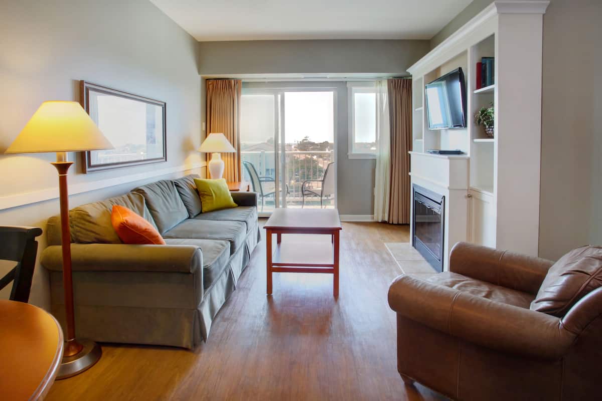 A modern living room with a sofa, a coffee table, and a leather armchair. There is a flat-screen TV mounted on a white cabinet, and a large window with curtains.