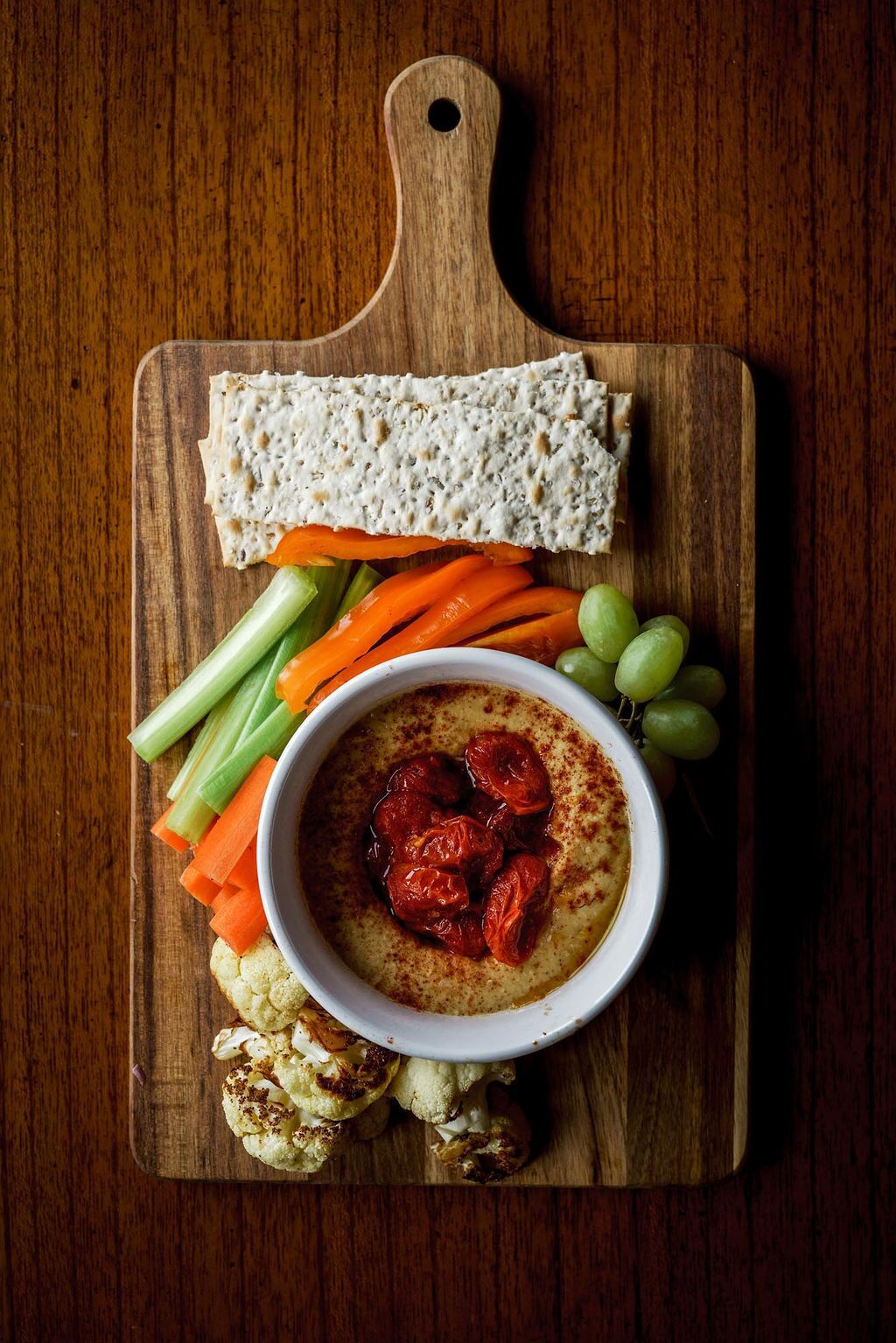 Una tabla de madera con un cuenco de salsa, zanahorias en rodajas, apio, coliflor, uvas y galletas.
