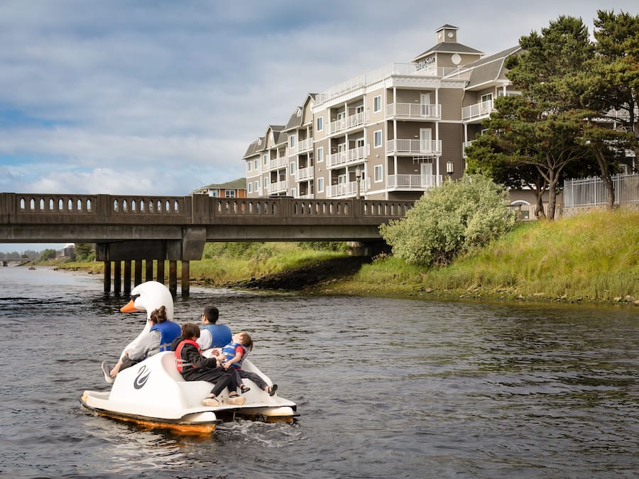 A family on a swan-shaped boat on a river, with a bridge and a building in the background.