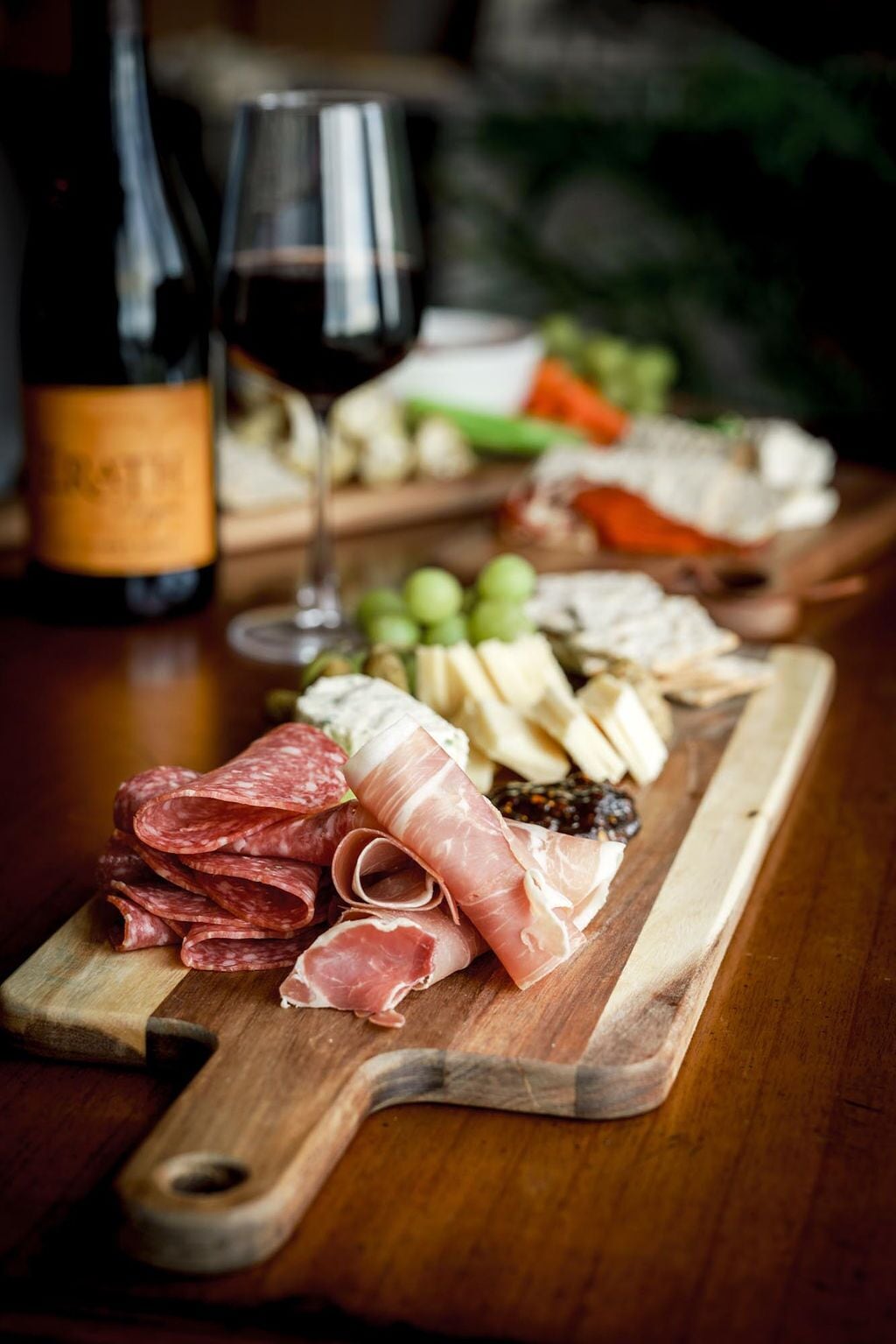 A wooden board with various cured meats, cheeses, and grapes, accompanied by a bottle of wine and a glass.