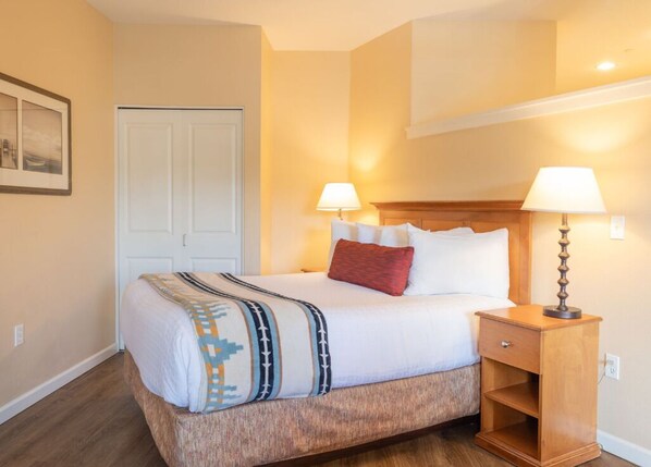 A hotel room with a bed, bedside lamps, a wooden nightstand, and a door. - Rivertide Suites Hotel (Seaside)