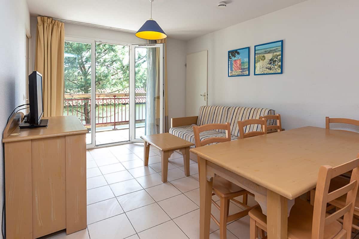 A room with a wooden dining table, chairs, a striped sofa, a TV on a wooden cabinet, and a sliding glass door leading to a balcony.