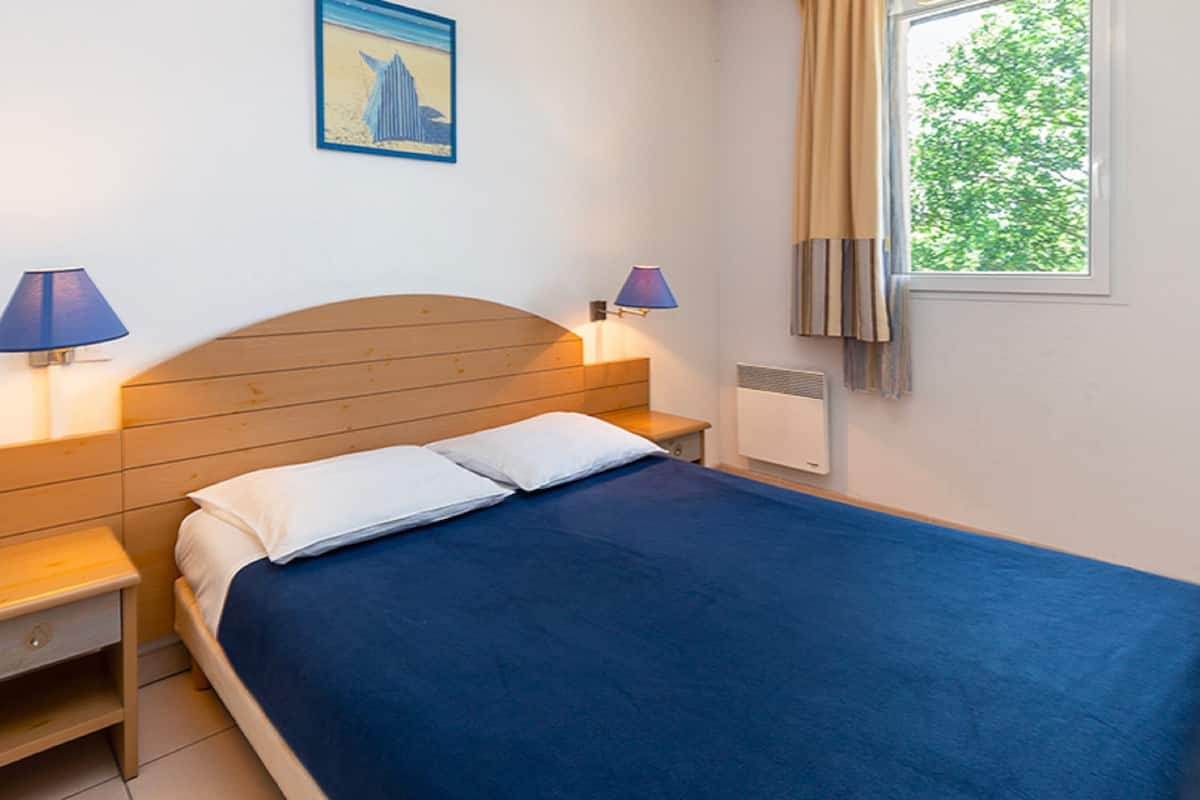 A bedroom with a wooden headboard, a blue bedspread, two bedside lamps, a nightstand, a radiator, and a window with curtains.