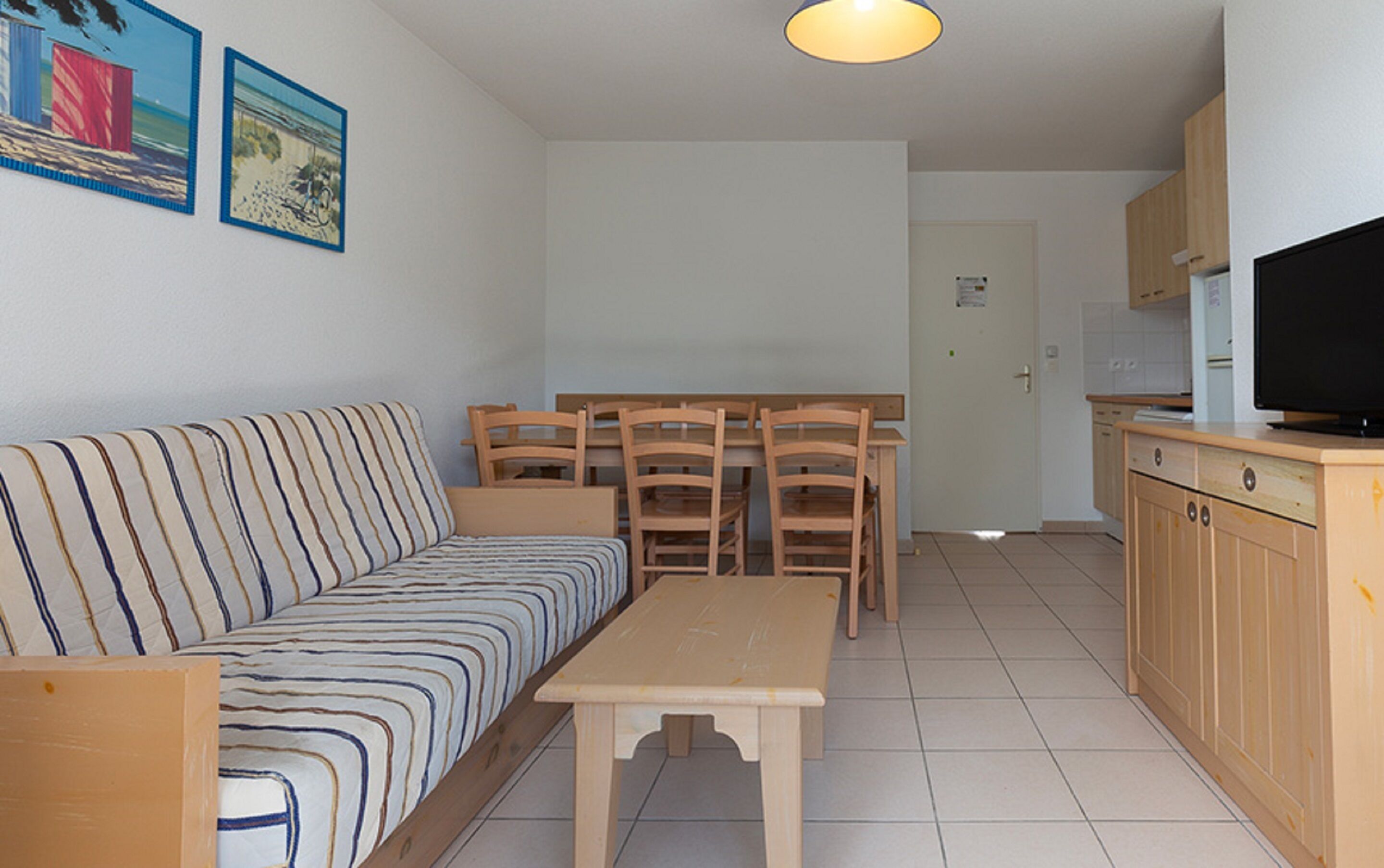 A compact living space with a sofa, a dining table, wooden chairs, a kitchen area with cabinets and a TV.