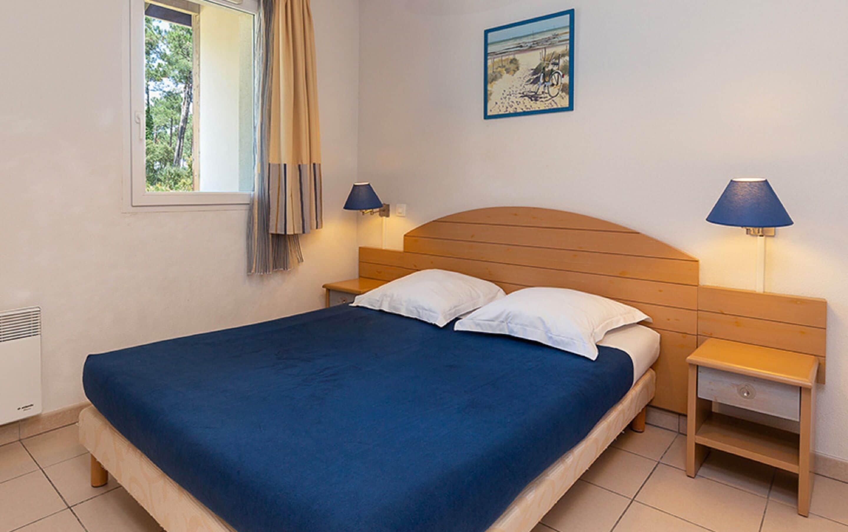 Une chambre avec un lit simple, une tête de lit en bois, deux tables de chevet avec des lampes, une fenêtre avec des rideaux et un tableau encadré au mur.