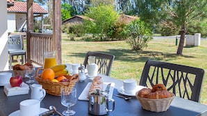 Montaje de desayuno al aire libre con una mesa, sillas y una variedad de alimentos, como frutas, pan y bebidas.