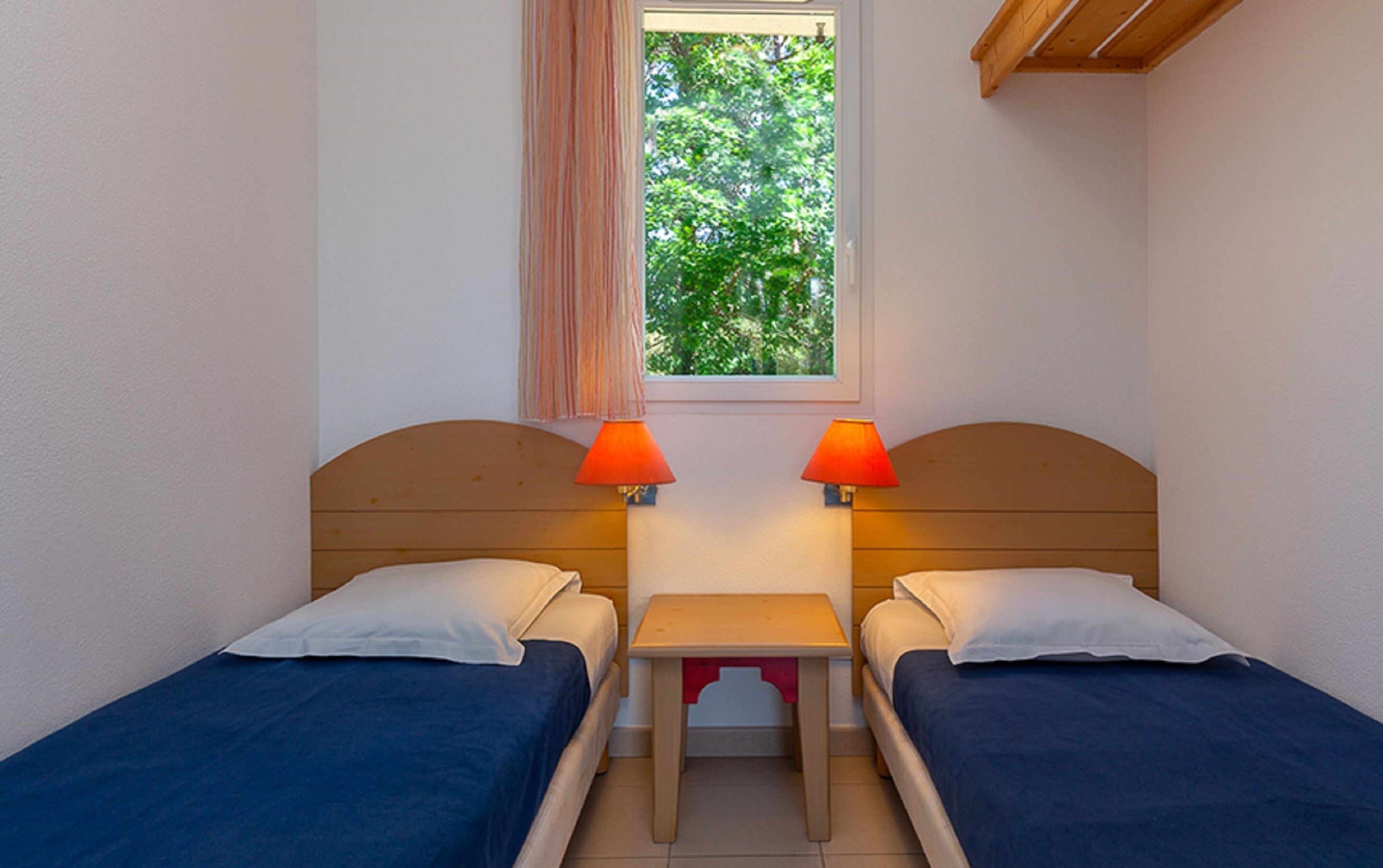 Een kleine kamer met twee eenpersoonsbedden, een tafeltje en twee lampen.