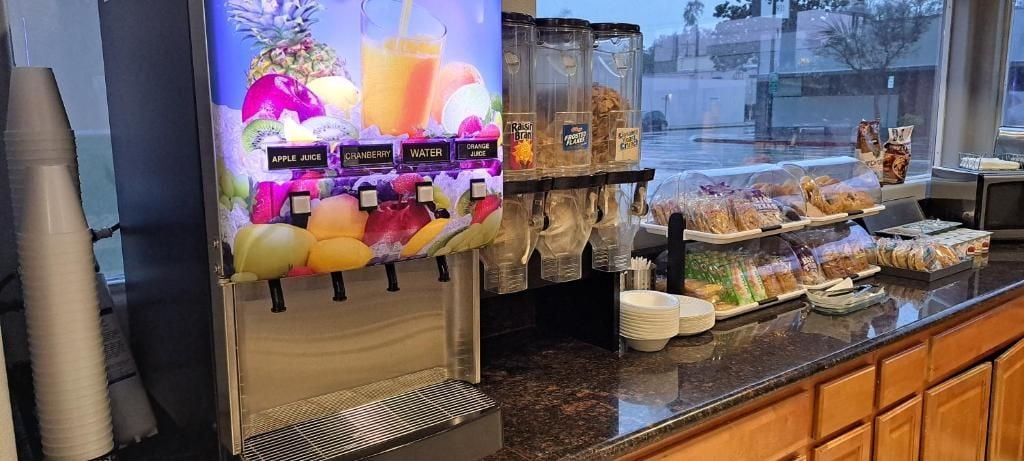 Un dispensador de zumos con varias opciones, una vitrina refrigerada con diversos alimentos y un mostrador con pan y bollería.