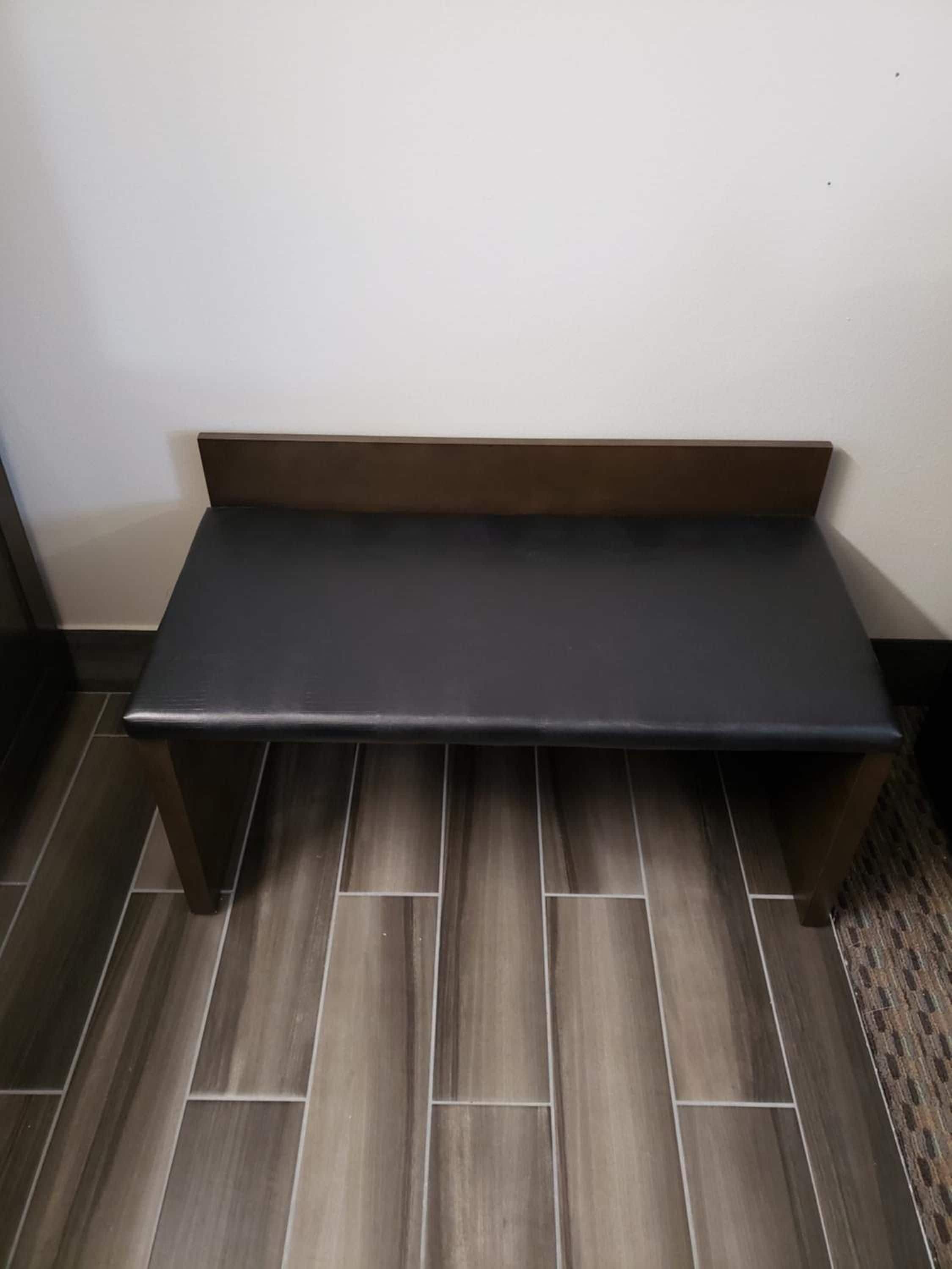 Un banco con superficie oscura y patas de madera, colocado frente a una pared con suelo de baldosas.