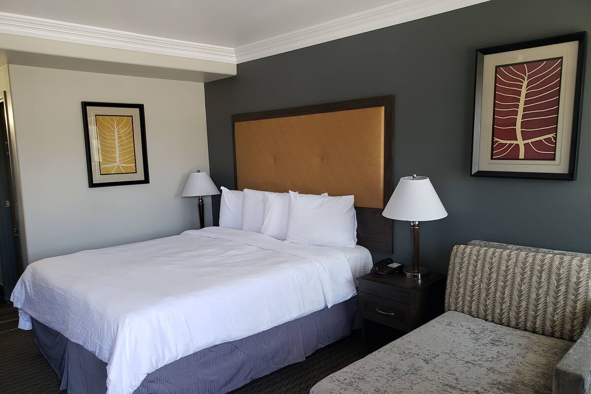 A hotel room with a large bed, two bedside tables with lamps, a patterned armchair, and two framed artworks on the walls.