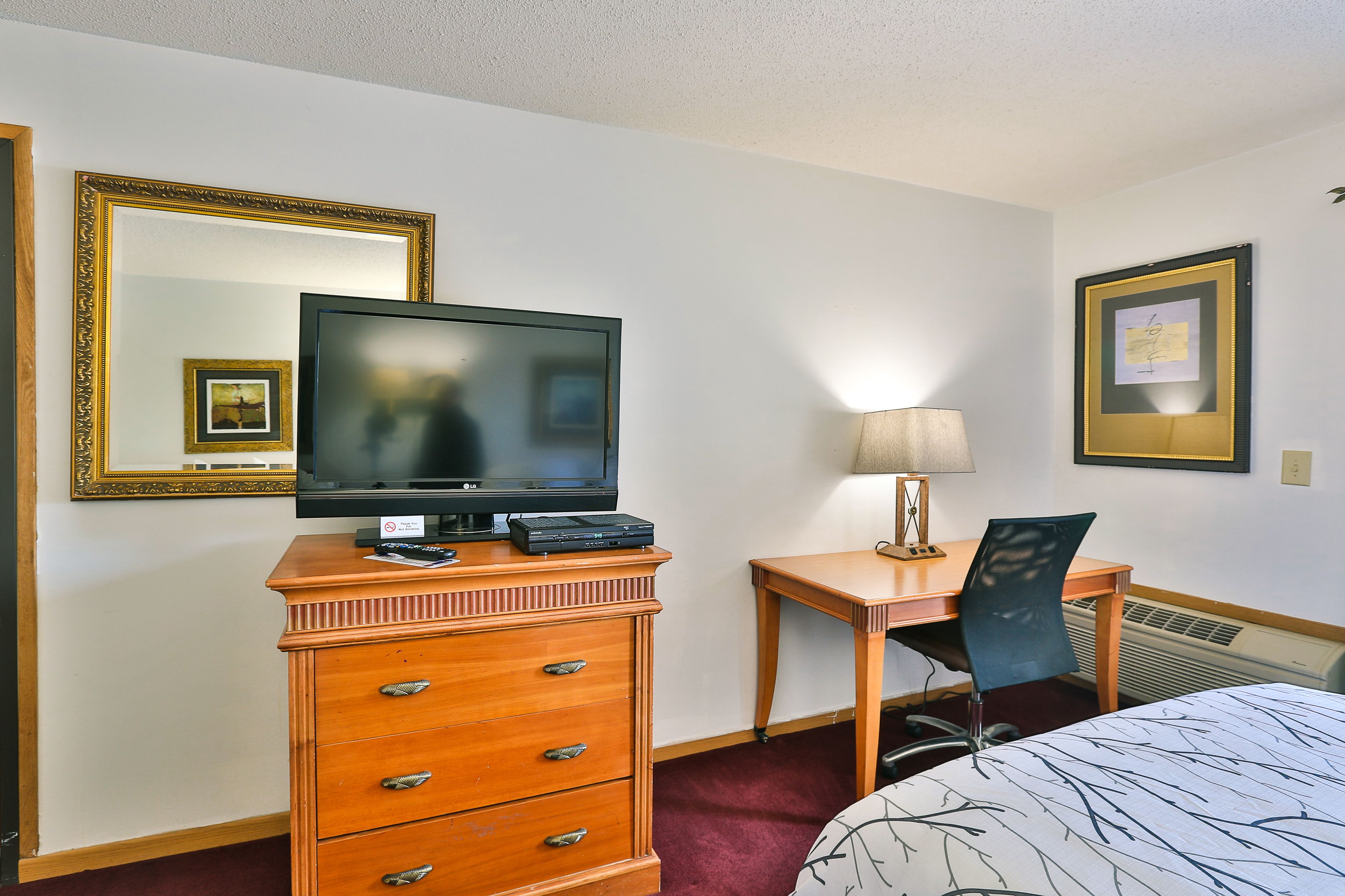 A hotel room with a wooden dresser, a television, a desk with a chair, a lamp, and a framed picture on the wall.