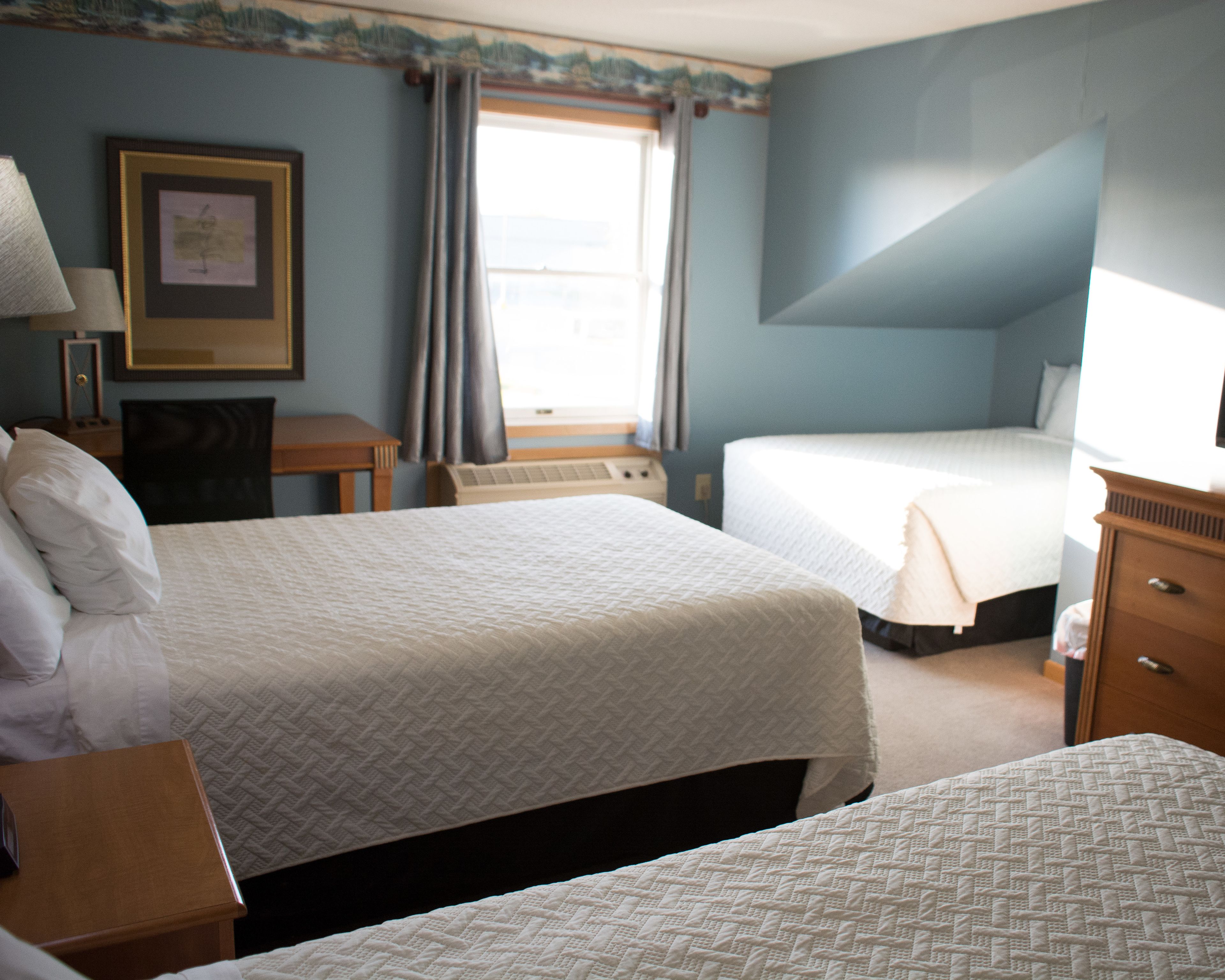 A hotel room with two beds, a desk, a chair, a lamp, a framed picture, and a window with curtains.