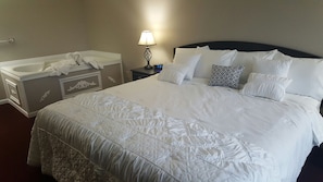 Habitación de hotel con una cama grande, una mesita de noche con lámpara, un tocador, un baño con bañera y un perchero para toallas.