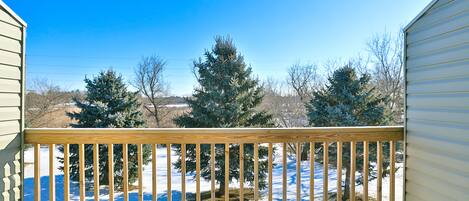 Una terraza de madera con barandilla, un patio cubierto de nieve y árboles de hoja perenne al fondo.