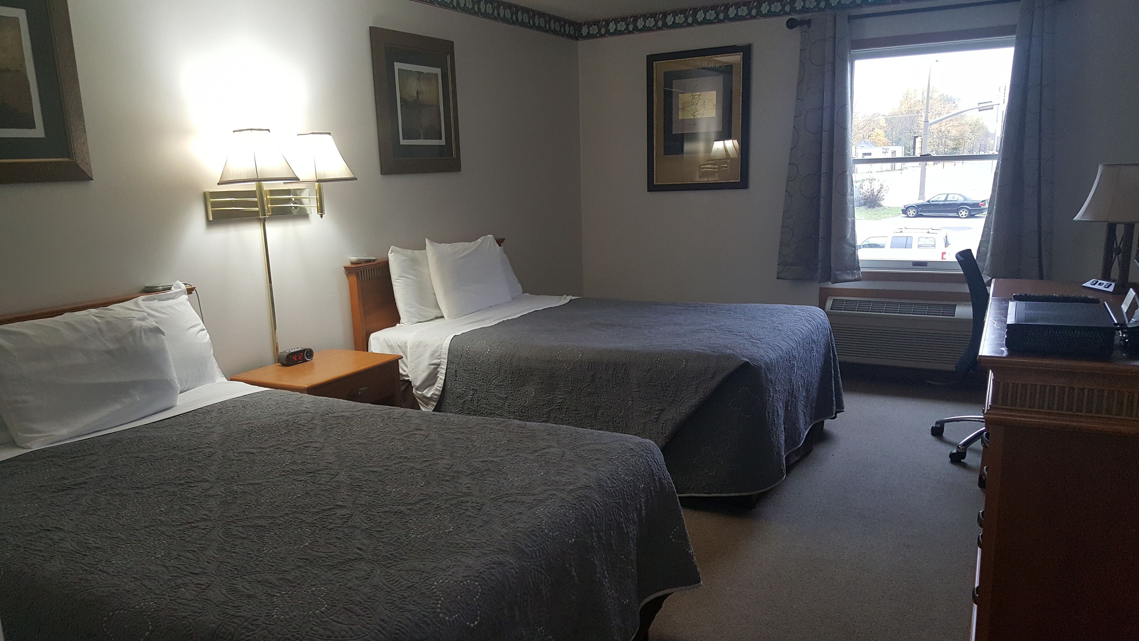 A hotel room with two beds, a desk, a lamp, and a window with a view of the street.