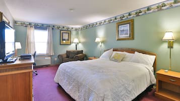 Ein Hotelzimmer mit einem großen Bett, zwei Nachttischlampen, einem Schreibtisch mit Computer, einem Sessel und einem Fenster mit Vorhängen.