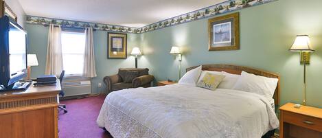 A hotel room with a large bed, two bedside lamps, a desk with a computer, a chair, and a window with curtains.