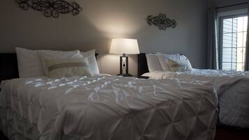 A hotel room with two beds, a bedside table with a lamp, decorative wall art, and a window with blinds.