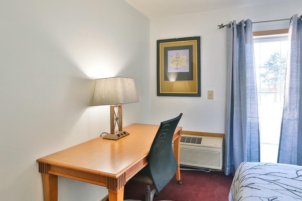 A hotel room with a wooden desk, a chair, a bed, a framed picture on the wall, and a window with curtains.