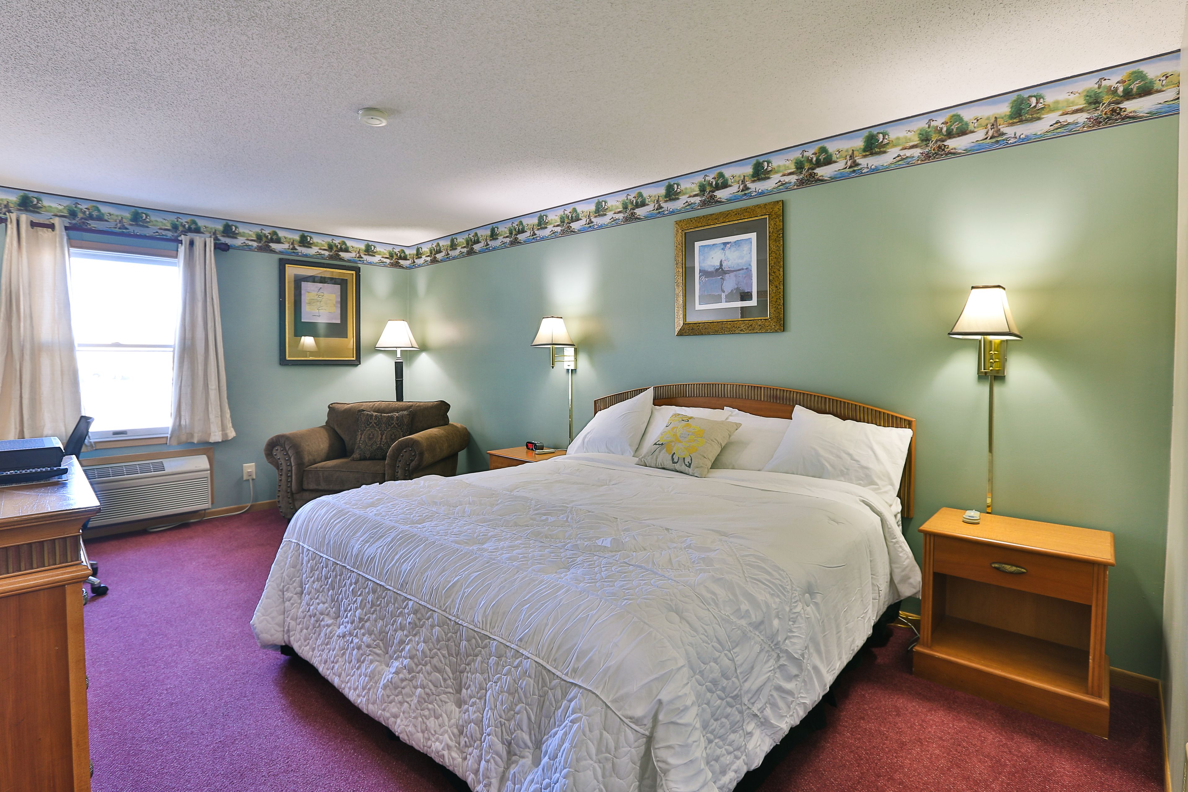 A bedroom with a large bed, two bedside lamps, a nightstand, a chair, a desk, and a window with curtains.
