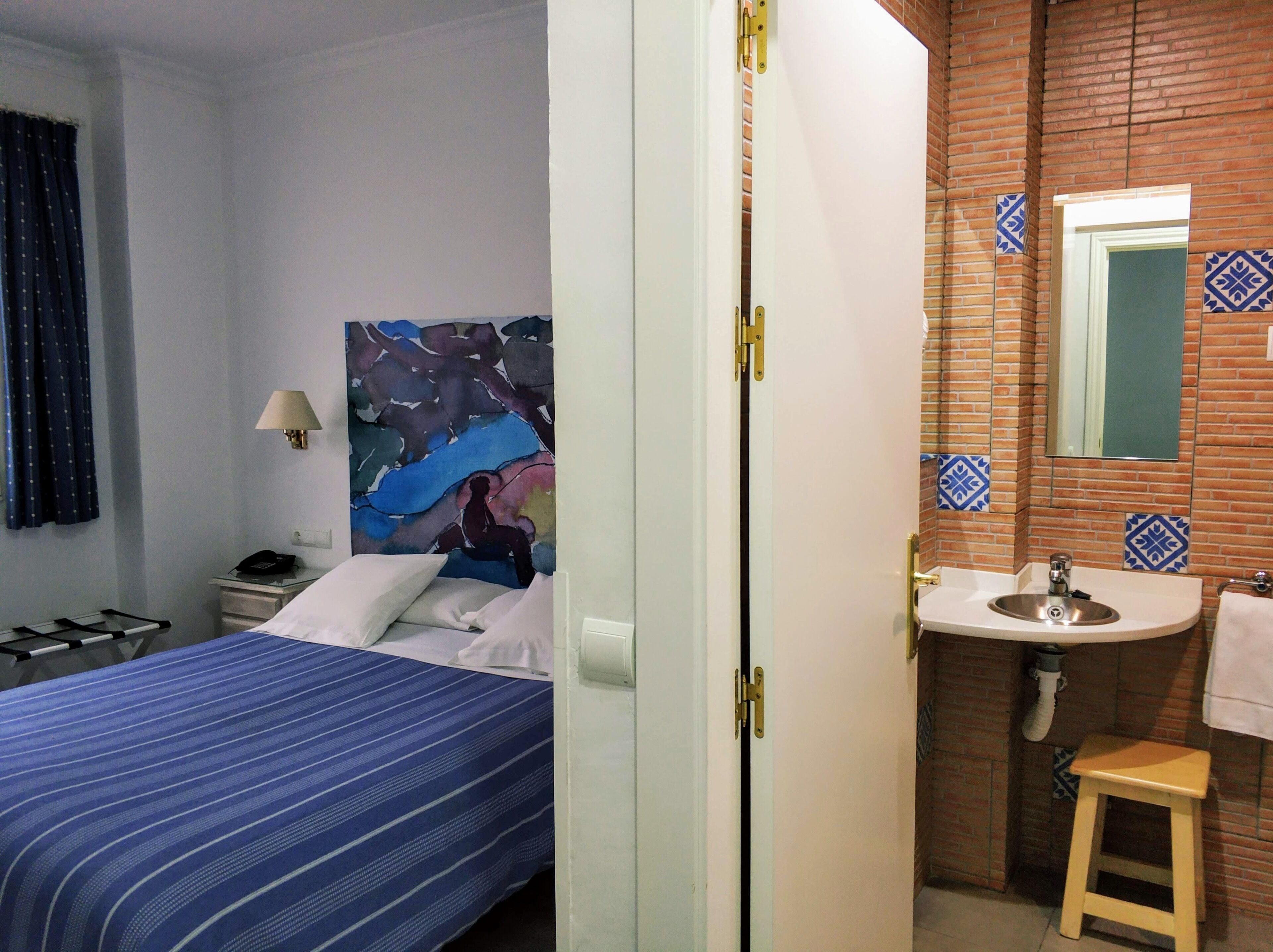 Hotelkamer met een bed, nachtkastje, lamp, schilderij, badkamer met wastafel en spiegel, en een klein krukje.