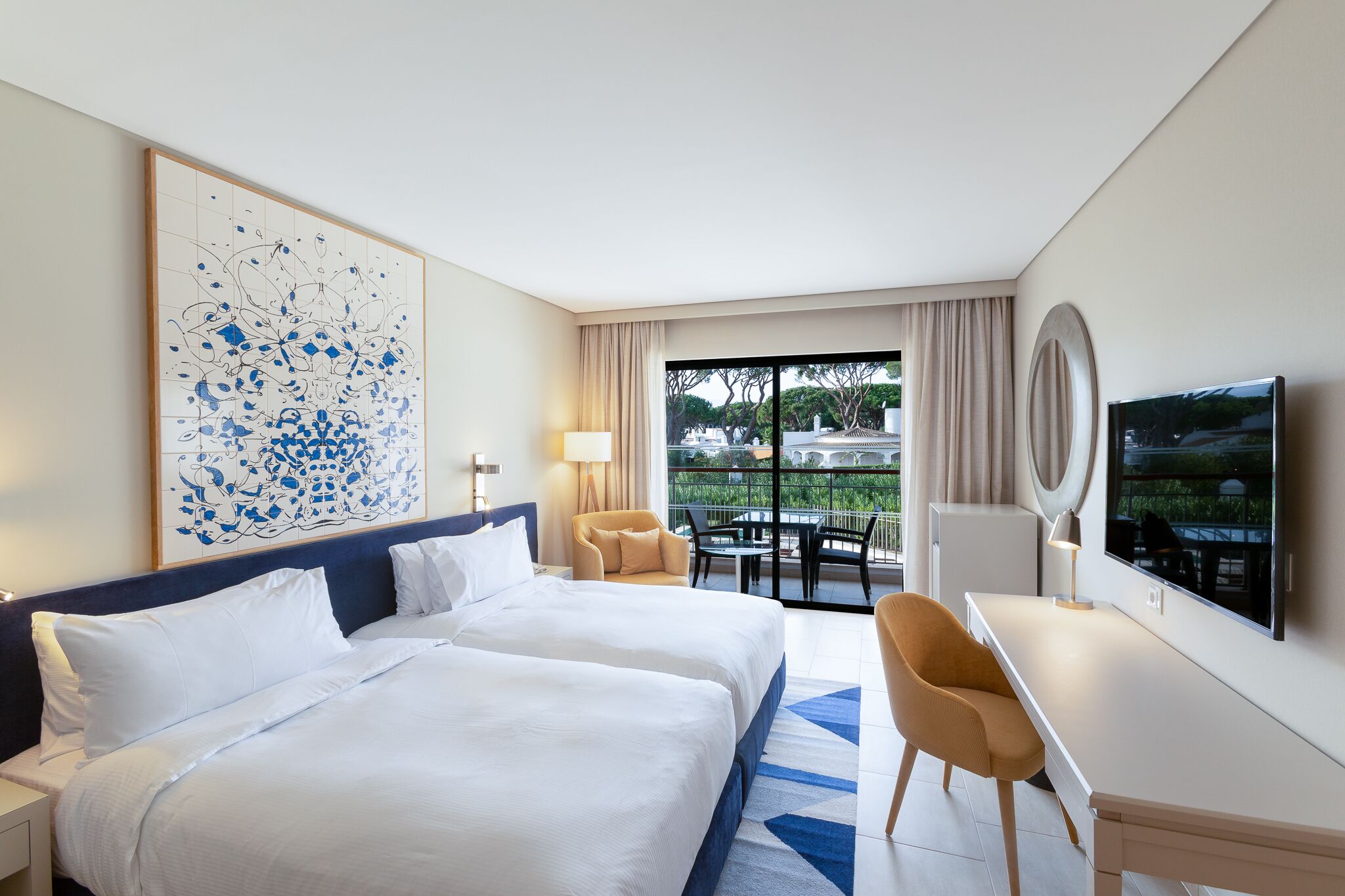 A hotel room with two beds, a desk, a TV, and a balcony with a view.