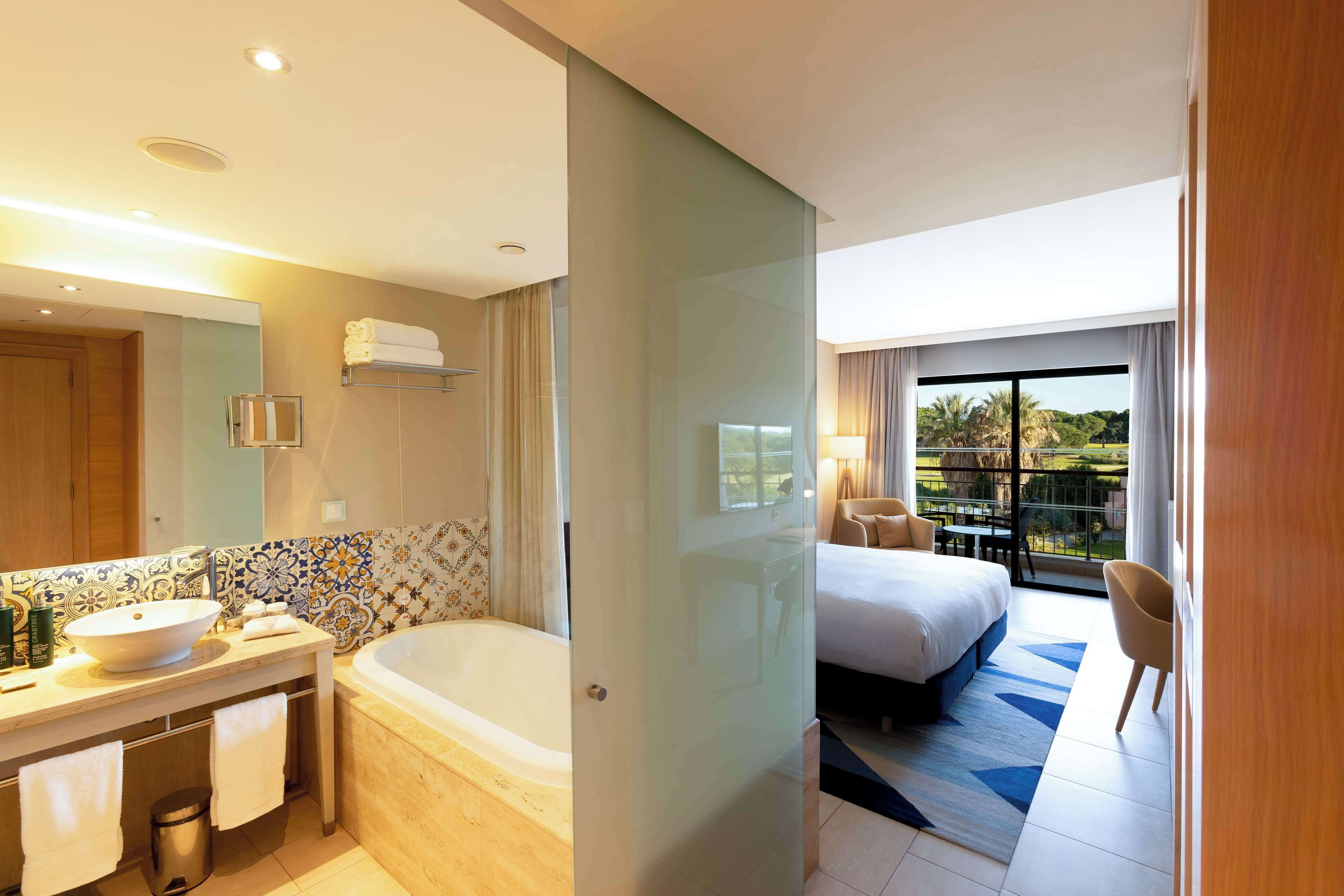 Ein modernes Hotelzimmer mit einer großzügigen Badewanne, separater Duschkabine, einem Balkon mit Tisch und Stühlen sowie einem großen Bett.
