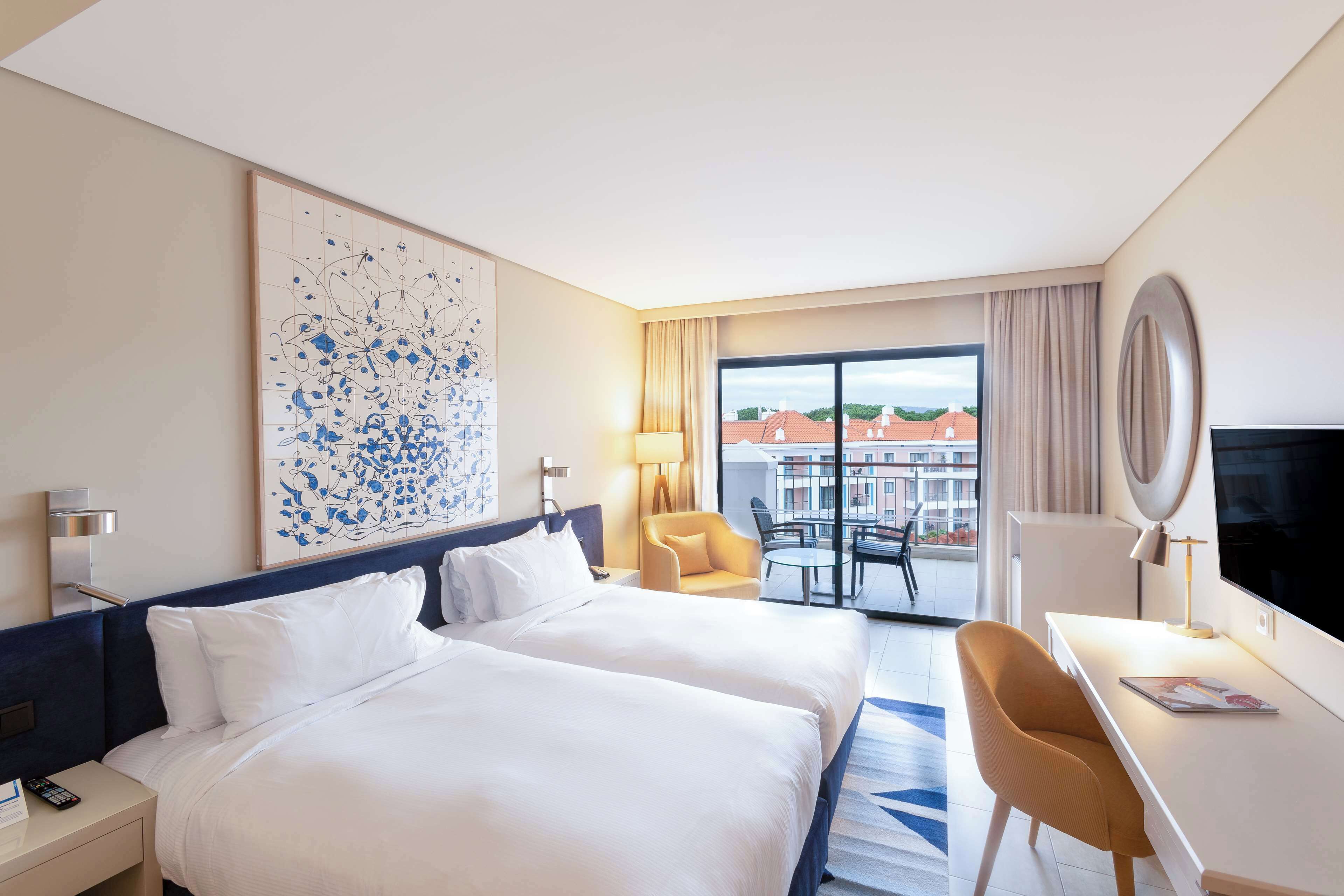Habitación de hotel con dos camas, un escritorio, una silla, un televisor y un ventanal con vistas a edificios.