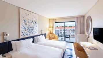 Habitación de hotel con dos camas, un escritorio, una silla, un televisor y un ventanal con vistas a edificios.