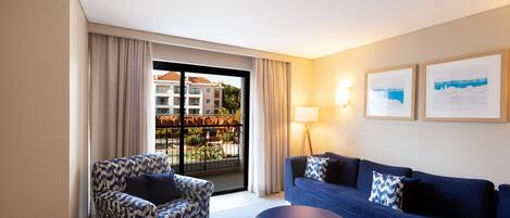 Habitación de hotel con sofá, sillón y una mesita. Tiene un balcón con vistas a edificios y zonas verdes.