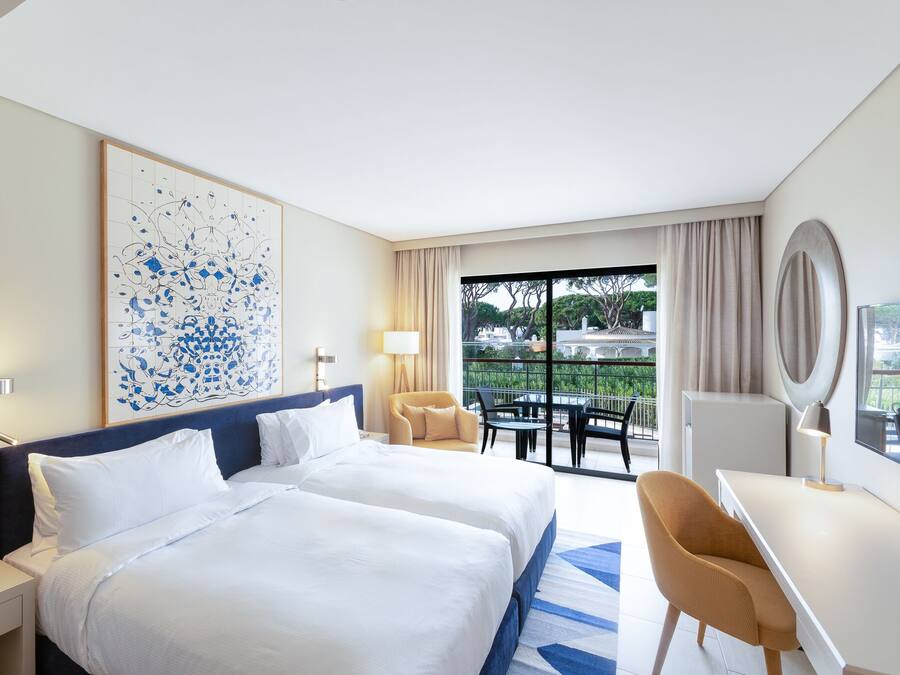 A hotel room with two beds, a desk, a chair, a balcony with a view, and a large abstract painting on the wall.