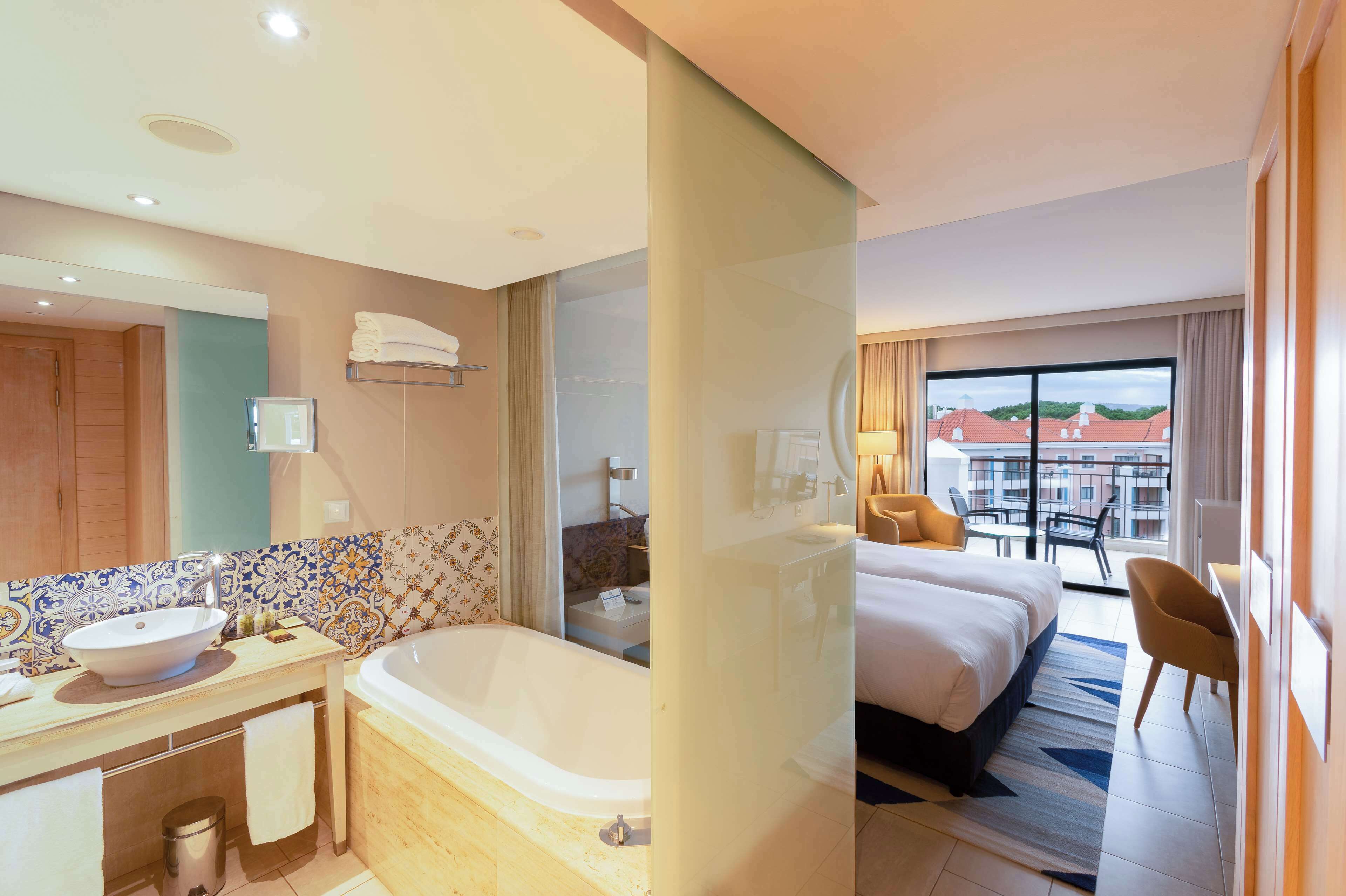 Une chambre d’hôtel dotée d’une grande baignoire, d’un lit double, d’un coin salon avec un fauteuil et offrant une vue sur les bâtiments par une grande fenêtre.
