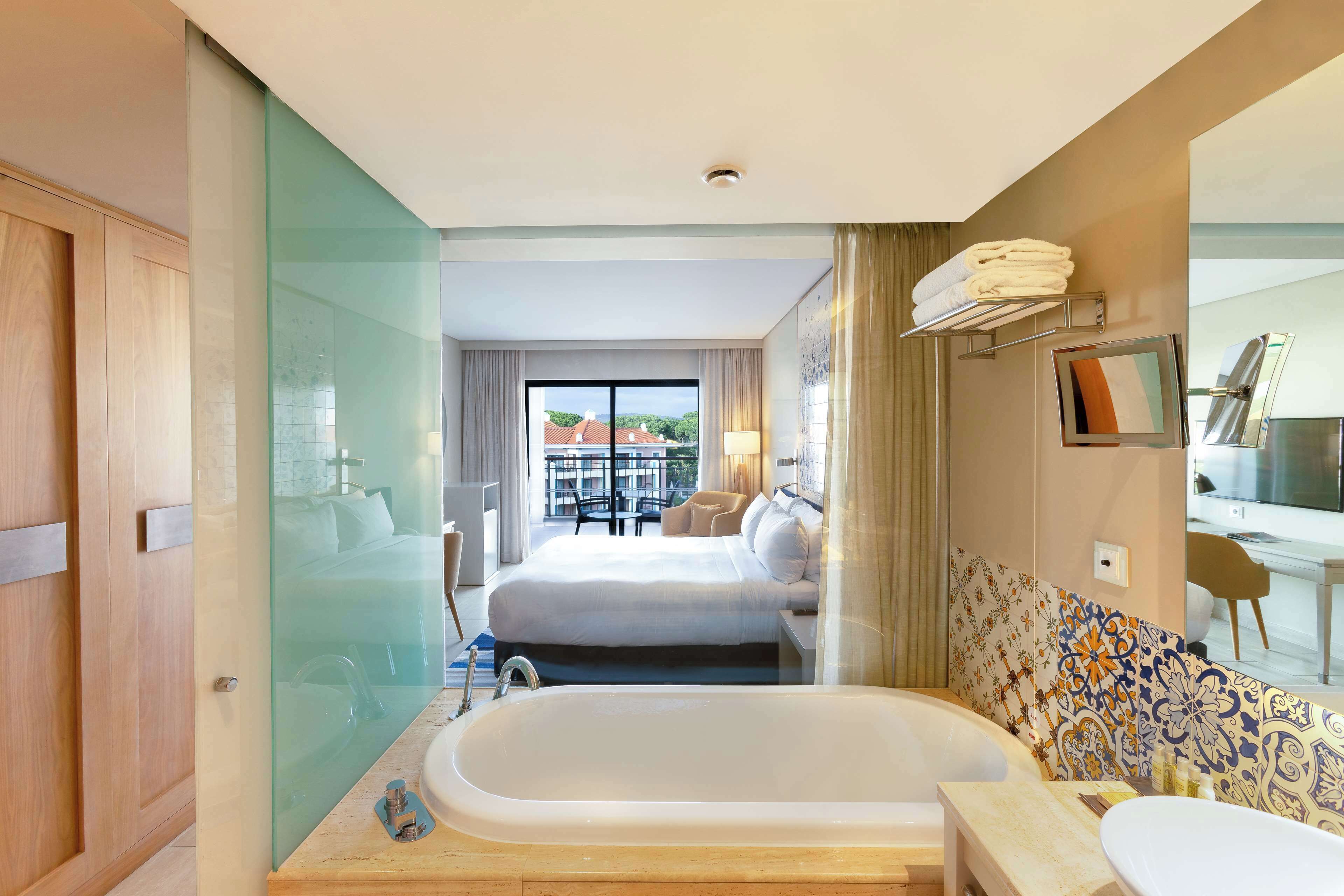 A modern hotel room with a large bathtub, a separate shower area, a bed with a view, and a dining area visible through the glass wall.