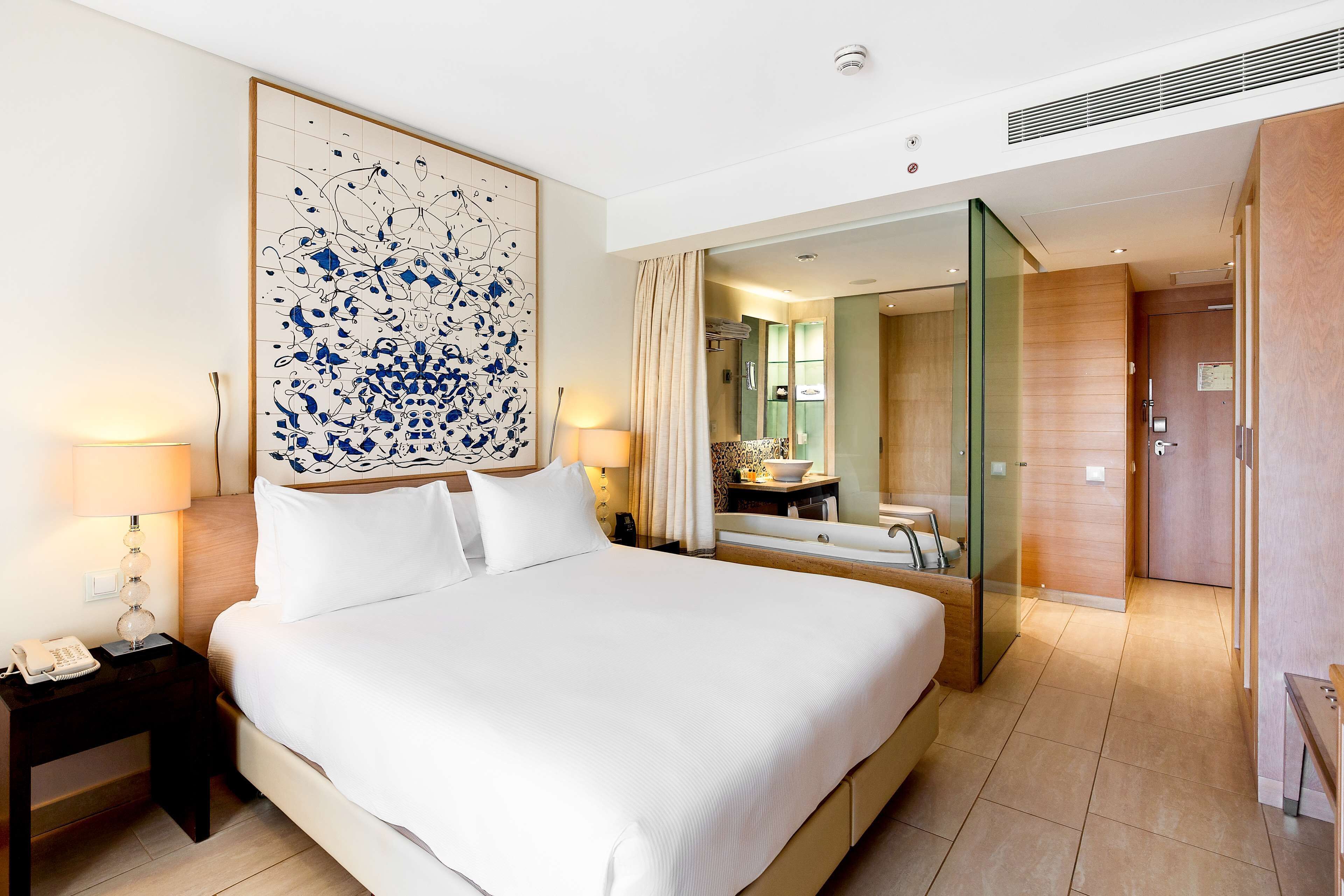 Une chambre d’hôtel avec un grand lit, des lampes de chevet, un téléphone, un miroir et une salle de bain visible à travers une porte en verre.