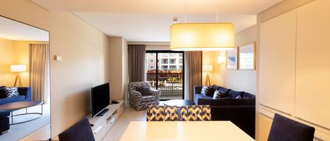 Habitación de hotel moderna con zona de comedor, sofá, televisor y vistas de la ciudad a través de amplias ventanas.