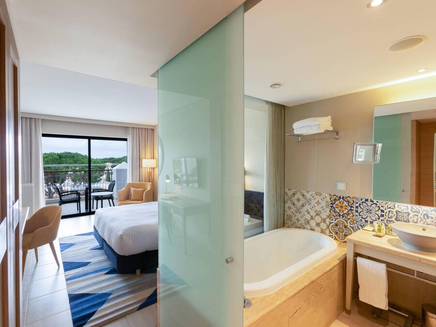 Hotelkamer met balkon, bed, bureau, stoel, ligbad en badkamer met wastafel en spiegel.