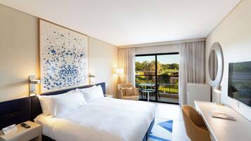 Quarto de hotel com cama grande, escrivaninha, cadeira, televisão e varanda com vista para o verde.