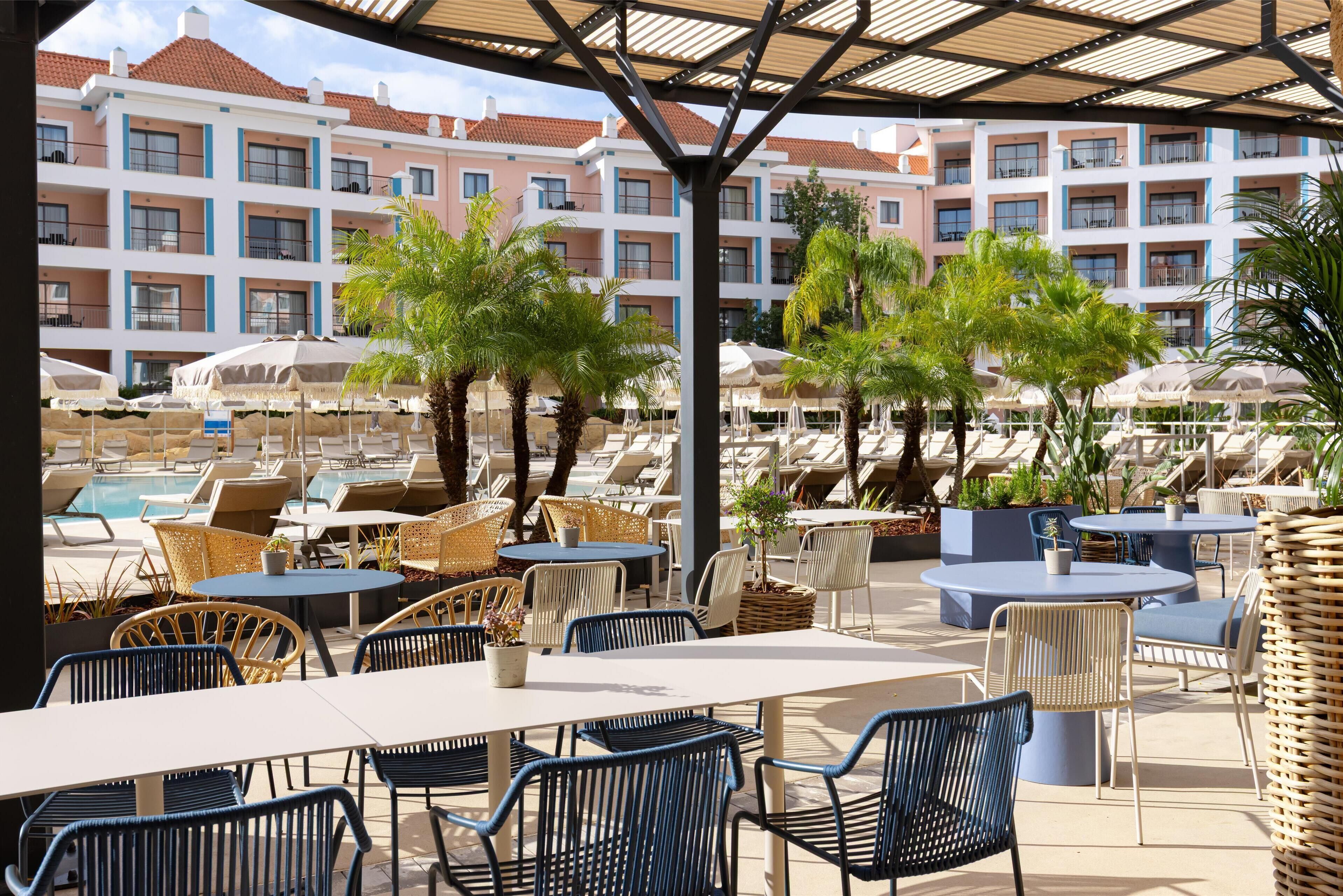 A resort pool area with lounge chairs and tables, surrounded by buildings with balconies.