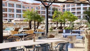 A resort pool area with lounge chairs and tables, surrounded by buildings with balconies.
