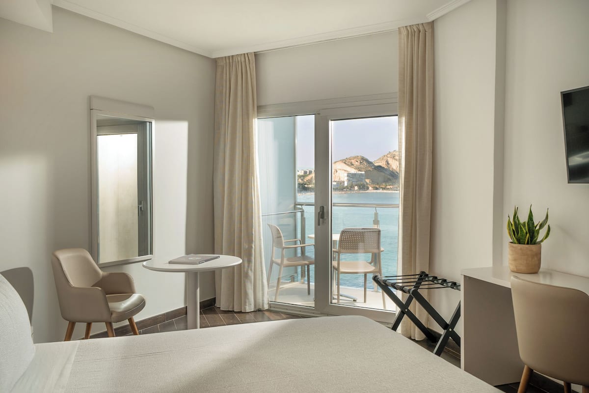 A hotel room with a bed, a desk, a chair, a small table, and a balcony with a view of the sea and mountains.