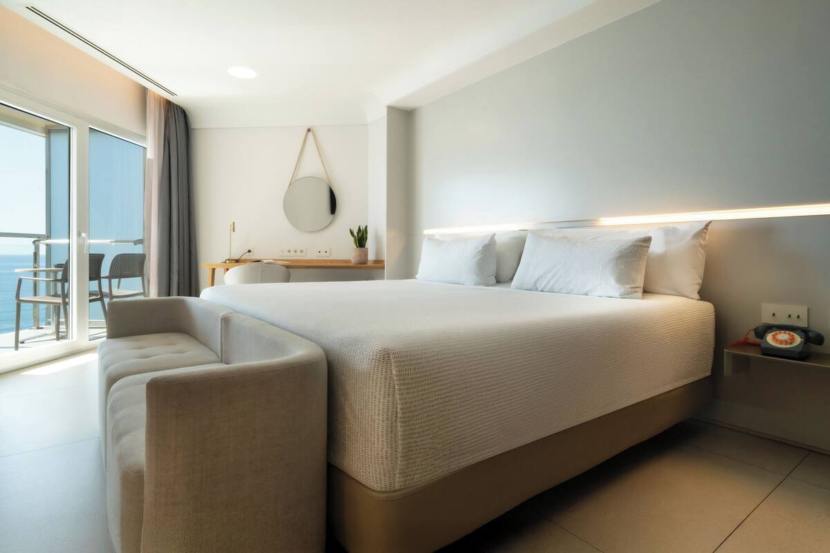 A modern hotel room with a large bed, a desk with a chair, a small table with a plant, and a view of the sea.