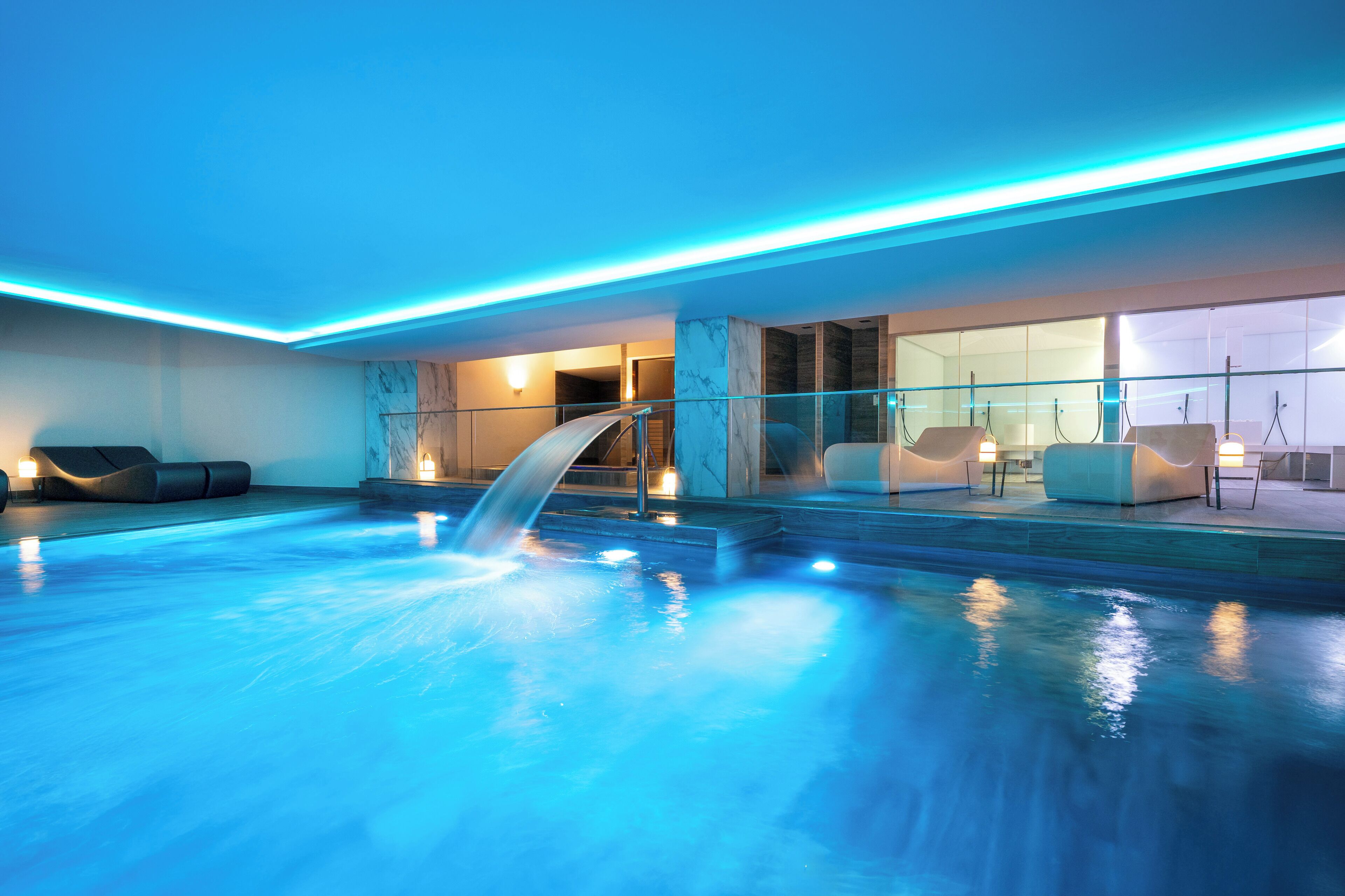 Una piscina interior moderna con caída de agua, barandillas de cristal y zonas de estar contemporáneas.