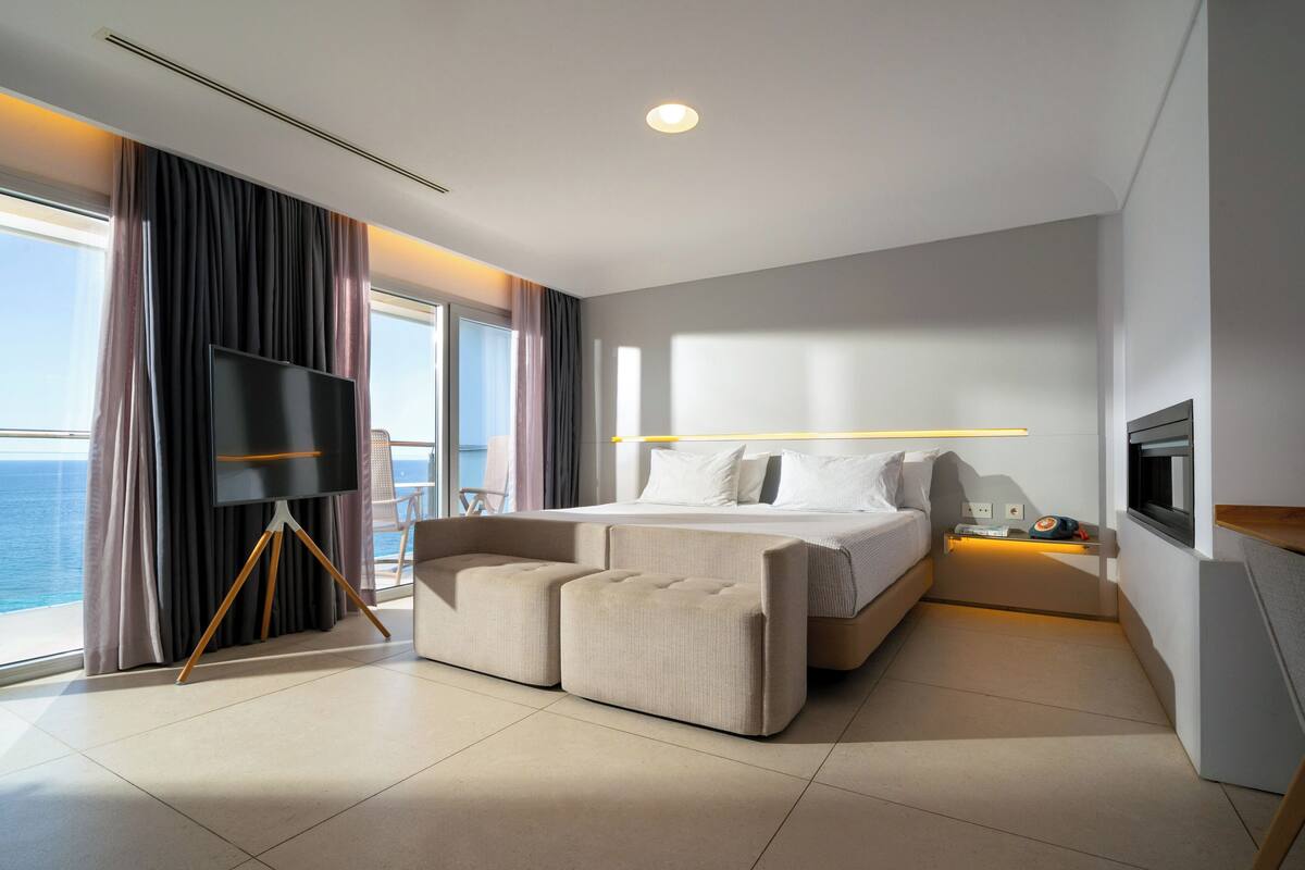 A modern hotel room with a large bed, a flat-screen TV, a seating area, and a view of the sea.