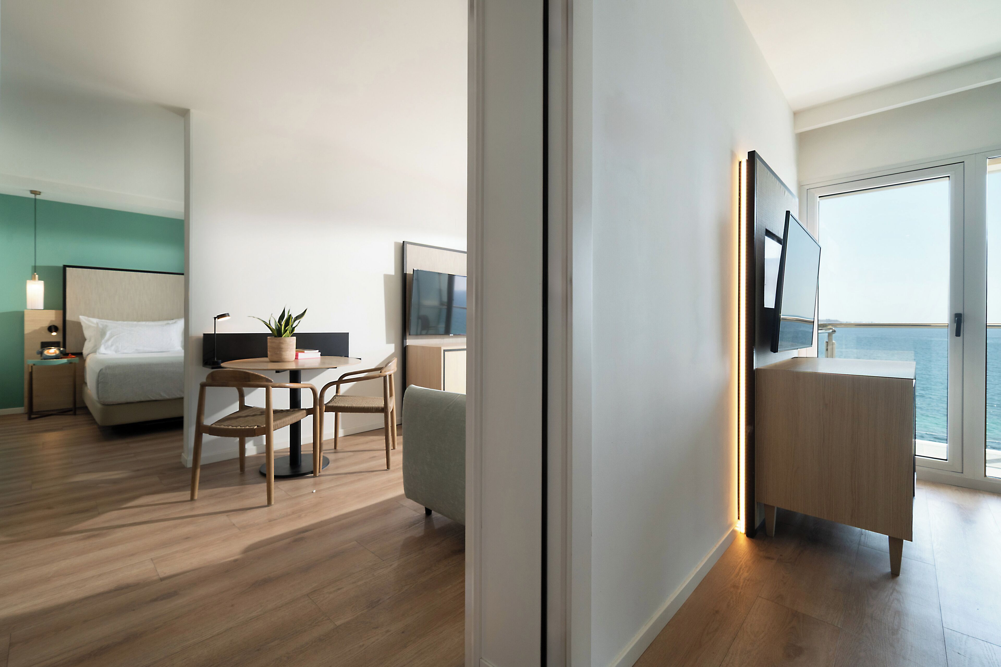 A modern hotel room with a bed, a dining area with a table and chairs, a desk with a TV, and a large window with a view of the sea.