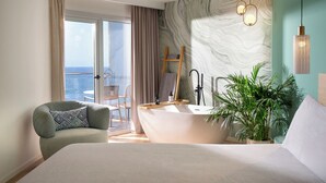 A modern hotel room with a large bed, a bathtub, a balcony with a view, and a potted plant.